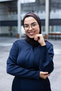 Vrolijk kijkende vrouw met hand onder haar kin. Ze draagt een bril, hijab en donkerblauwe blouse. Ze staat op een plein. 